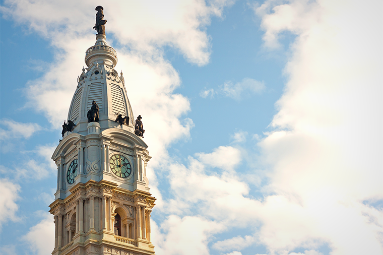A photo of the William Penn statue on top of Philadelphia's City Hall set against a cloudy blue sky