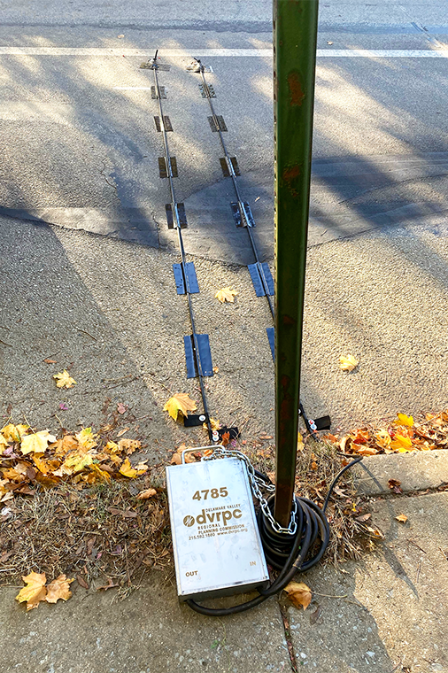 A photo of a DVRPC branded traffic count box chained to a street pole, with the sensors extending across a lane of a road
