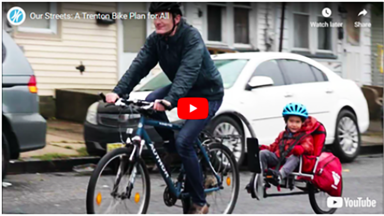 A screenshot of a video for "Our Streets: A Trenton Bike Plan for All," which shows a man riding a bike with a child riding in a cart being pulled by the bike.