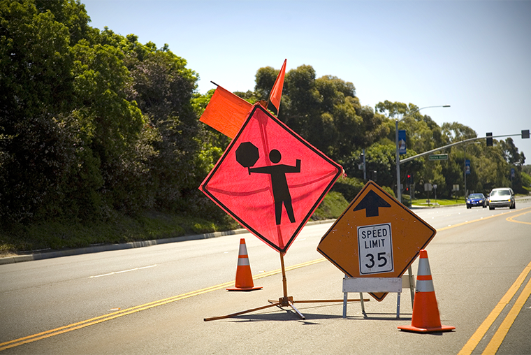 A photo of construction signs in a road signaling a reduced speed limit and a person directing traffic ahead
