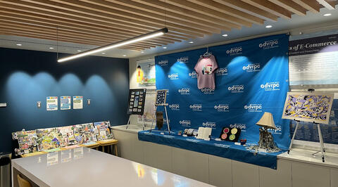 A corner of the DVRPC lobby set up as a small art gallery displaying staff artwork. One side features the collaborative collage, while the other displays nine handmade items including textiles, greeting cards, and a book of poetry.
